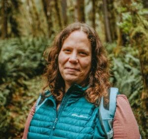 Image Description: A headshot of Syren Nagakyrie, a white nonbinary person with wavy brown hair. They are wearing a backpack and outdoor clothing. A blurry mossy green forest is in the background.